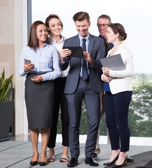 Group of five cheerful business people standing in office lobby, looking at digital tablet computer in his young leaders hands and smiling